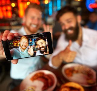 Bearded businessmen having a lunch meeting in the restaurant and posing for a selfie image. Drinks and food are on the table.