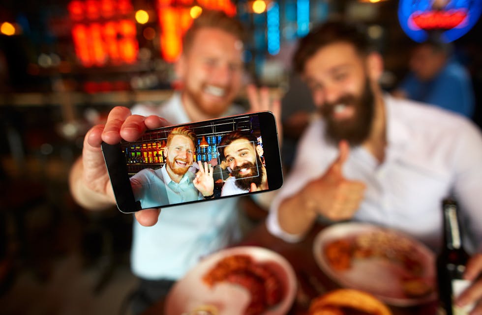 Bearded businessmen having a lunch meeting in the restaurant and posing for a selfie image. Drinks and food are on the table.